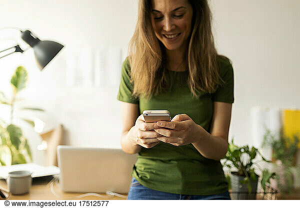 Smiling young woman using smart phone while standing at desk in home office