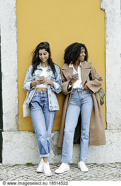 Smiling young woman using smart phone with friend leaning on yellow wall