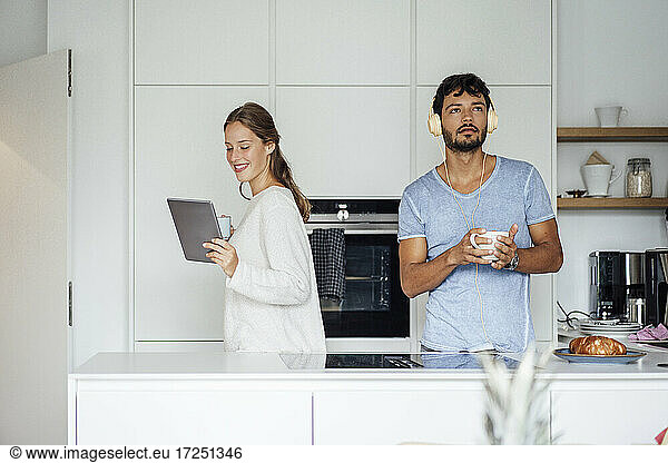Smiling young woman using tablet while man listening music in kitchen