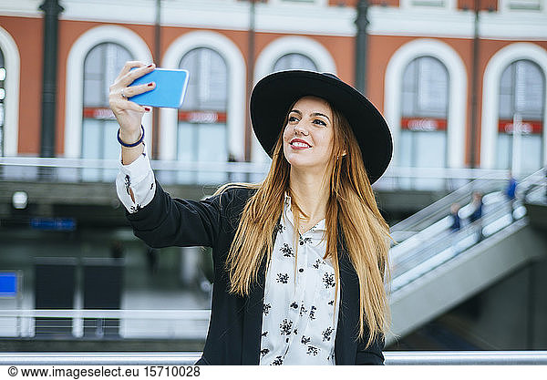 Smiling young woman wearing a hat taking a selfie at train station