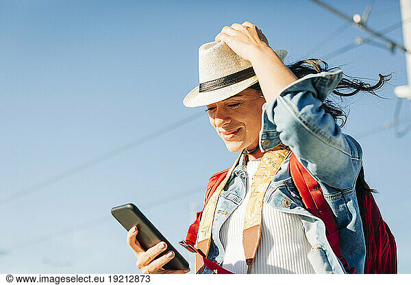 Smiling young woman wearing hat using smart phone under sky