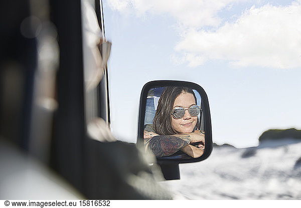 Smiling young woman wearing sunglasses in side-view mirror of car