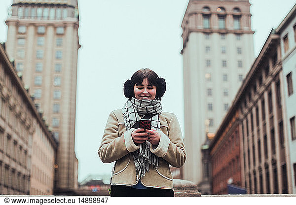 Smiling young woman wearing warm clothing while using mobile phone against buildings in city
