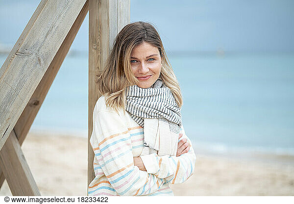 Smiling young woman with arms crossed leaning on lifeguard hut at beach