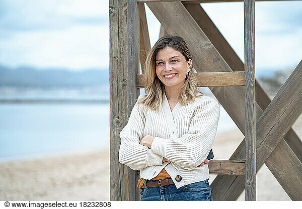 Smiling young woman with arms crossed leaning on wooden lifeguard hut at beach
