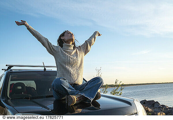 Smiling young woman with arms raised sitting on car hood