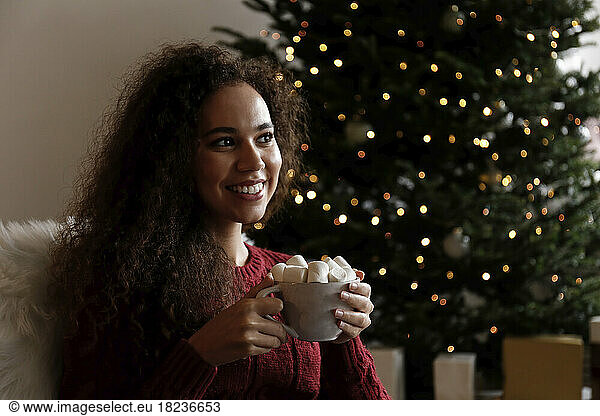 Smiling young woman with cup of marshmallow cocoa sitting in front of Christmas tree