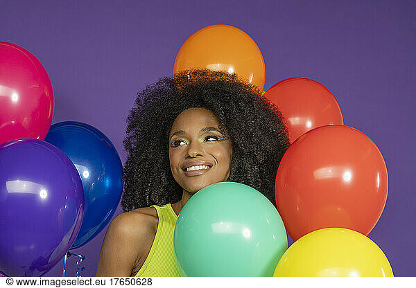 Smiling young woman with curly hair amidst colorful balloons against purple background