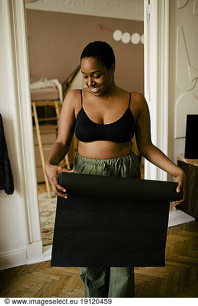 Smiling young woman with exercise mat standing at home