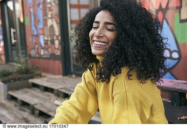 Smiling young woman with eyes closed sitting against wall in city