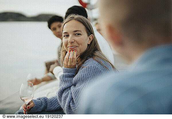 Smiling young woman with hand on chin sitting with friends on pier near lake