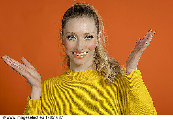 Smiling young woman with hand raised standing against orange background