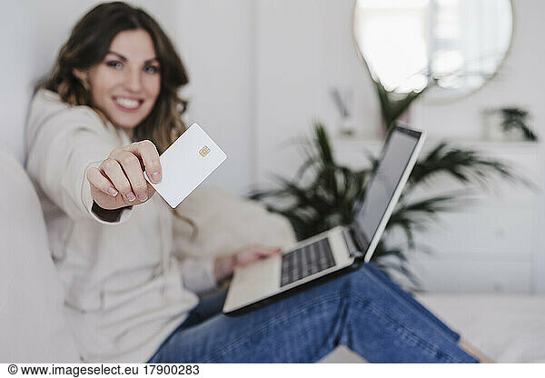 Smiling young woman with laptop showing credit card