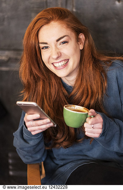 Smiling young woman with long red hair sitting at table  holding mobile phone and cup of coffee  looking at camera.