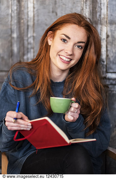 Smiling young woman with long red hair sitting at table  holding notebook and cup of coffee  looking at camera.