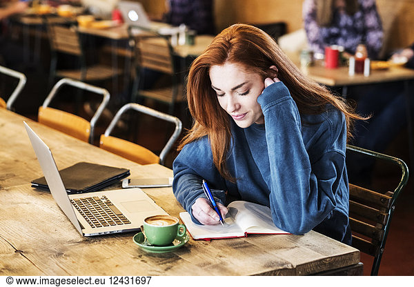 Smiling young woman with long red hair sitting at table  working on laptop computer.