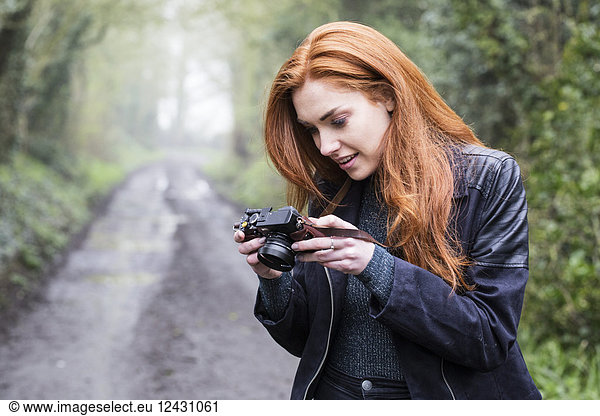 Smiling young woman with long red hair walking along forest path  taking pictures with vintage camera.