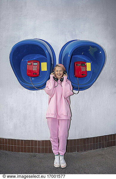 Smiling young woman with pink hair wearing pink hooded shirt standing in front of telephone booths