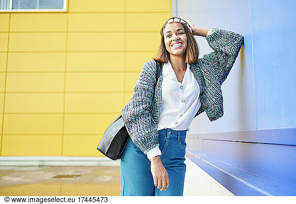 Smiling young woman with shoulder bag leaning on blue wall