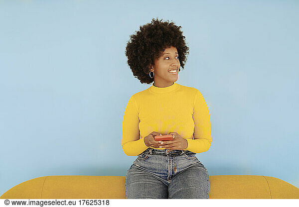 Smiling young woman with smart phone sitting on yellow sofa against blue background