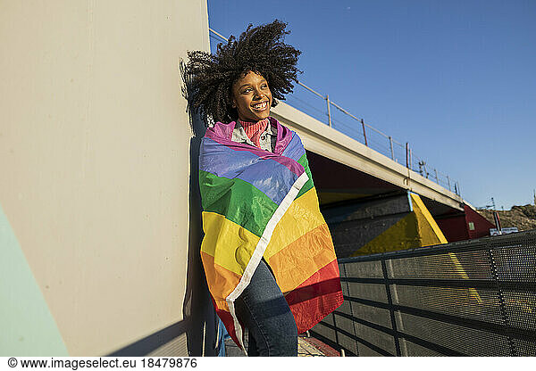 Smiling young woman wrapped in rainbow flag leaning on wall