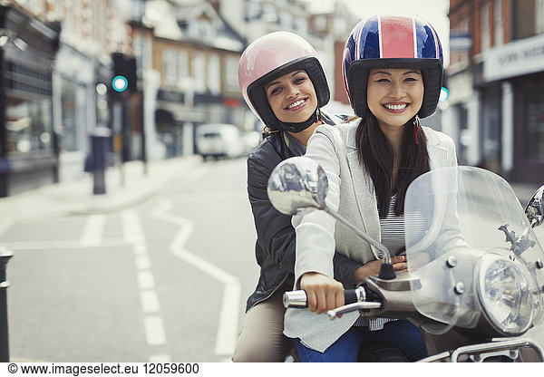 Smiling young women friends wearing helmets  riding motor scooter on urban street