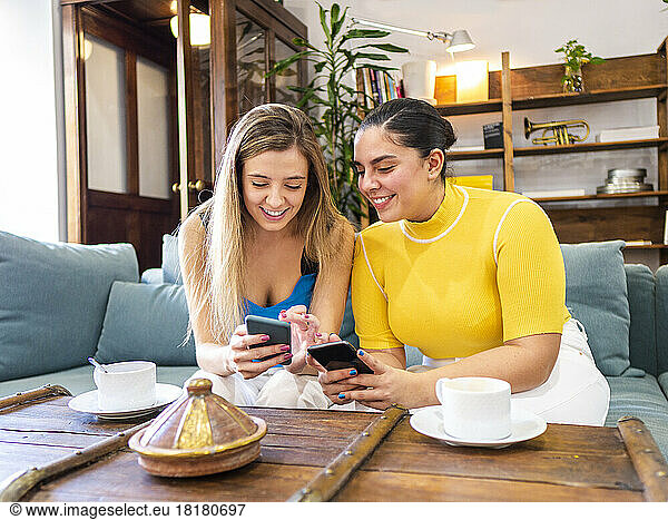 Smiling young women sharing mobile phones on sofa at home
