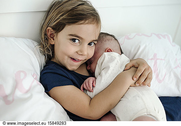 Smiling 4 yr old leaning against pillows holding newborn baby