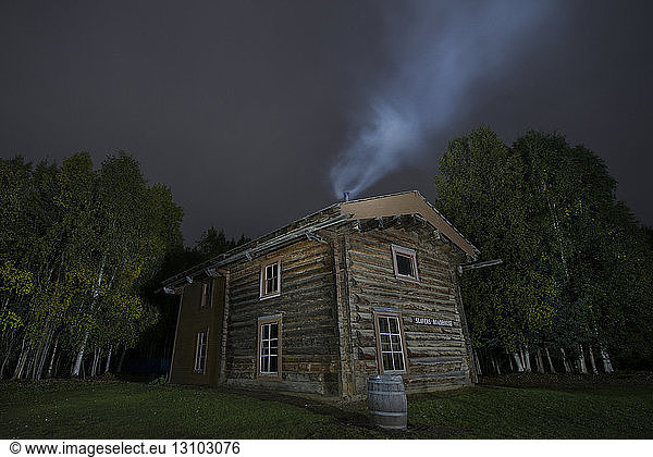 Smoke emitting from chimney on Slaven's roadhouse by trees at Yukon_Charley Rivers National Preserve against sky at night