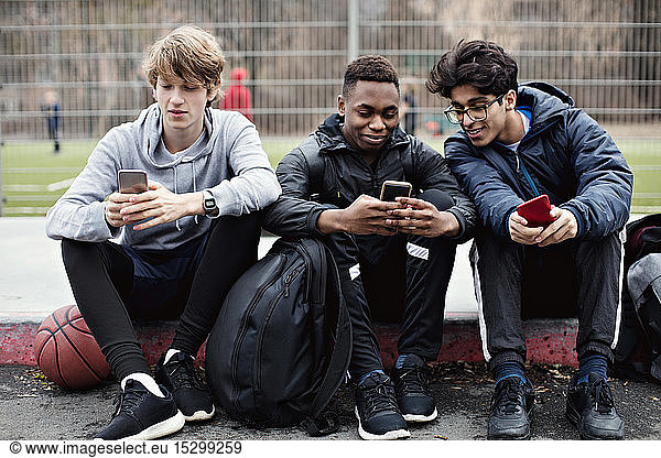 Social media addicted friends using mobile phones while sitting on sidewalk after basketball practice in city
