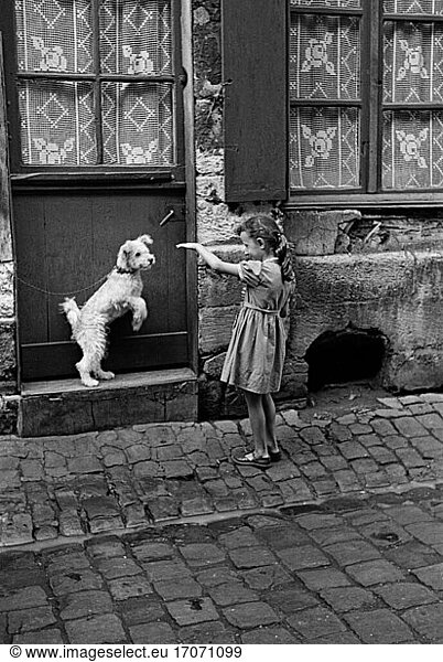 Society: Childhood.
– A girl playes with a little dog tied next to a doorway in Rouen  France.
Photo  1956.