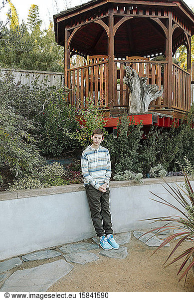 Solemn teen boy wearing blue shoes leaning on wall near gazebo