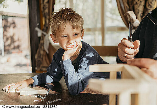 Son leaning on elbow looking at father making birdhouse at home