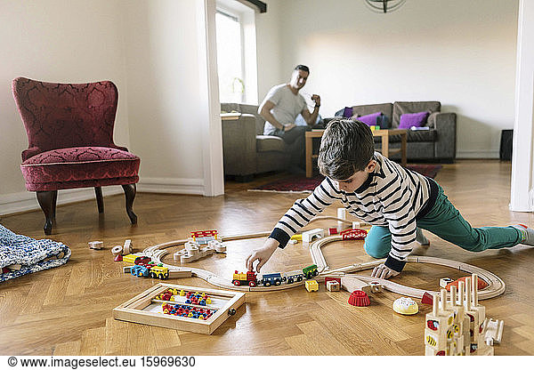 Son playing with miniature train while father sitting on sofa in background at home