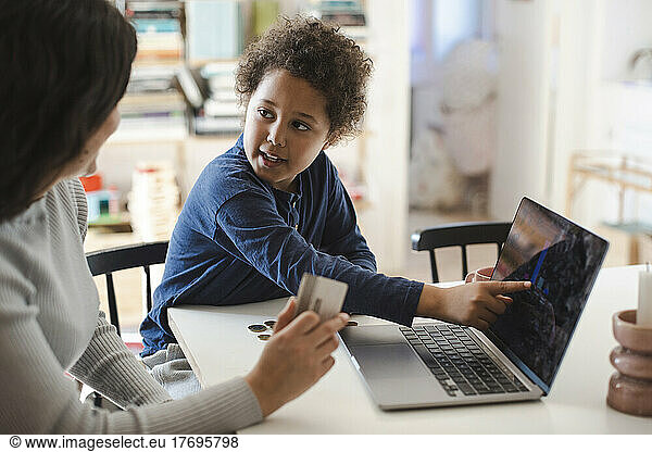Son pointing at laptop screen while looking at mother holding credit card