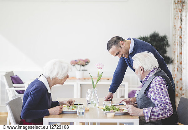 Son showing mobile phone to mother and father having lunch at table in nursing home