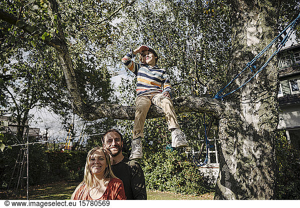 Son sitting on tree in garden with parents watching him