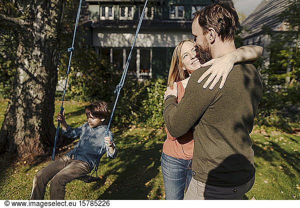Son swinging in garden with parents kissing next to him