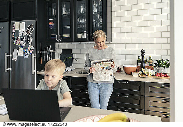 Son using laptop while mother reading newspaper in kitchen