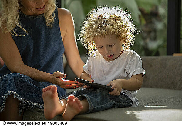 Son with curly hair using tablet PC sitting next to mother at home