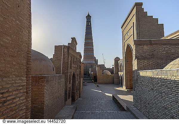 Sonnenaufgang  eine schmale Straße mit Backsteingebäuden und Blick auf ein hohes  aus runden Backsteinen erbautes Minarett in Chiwa  Usbekistan.
