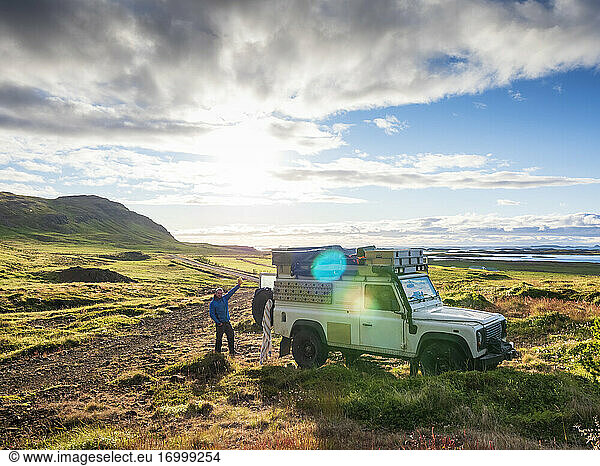 Sonnenuntergang über einem einsamen Reisenden  der hinter einem Geländewagen steht und der Kamera zuwinkt  Island