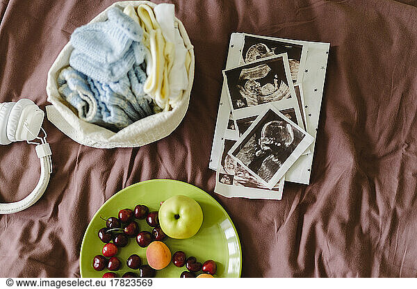 Sonograms with baby clothing and fresh fruits on bed