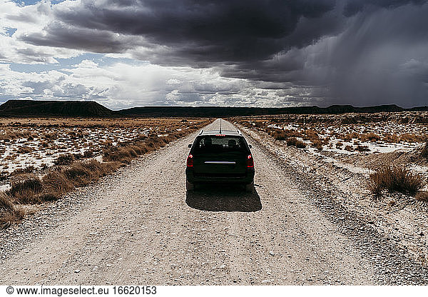 Spain  Navarre  Storm clouds over car driving along empty dirt road in Bardenas Reales