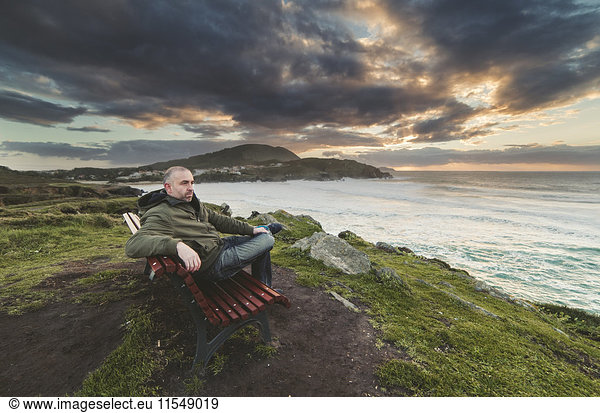 Spanien  Ferrol  Mann sitzt auf einer Bank an der Küste und schaut in die Ferne.
