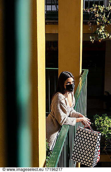 Spanish brunette girl with face mask leaning on a balustrade and holding shopping bags in a shopping mall. Vertical photo.