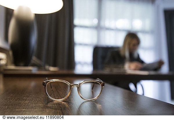 Spectacles on desk with businesswoman checking documents in the background