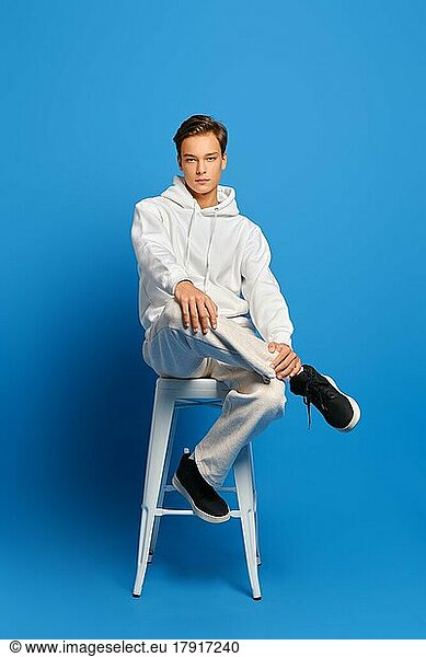 Sportlicher Mann in weißem Sweatshirt und Jogginghose sitzt auf einem hohen Stuhl vor blauem Studiohintergrund