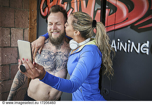Sportswoman kissing male athlete while taking selfie on smart phone against graffiti door