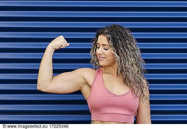 Sportswoman smiling while looking at bicep in front of corrugated wall